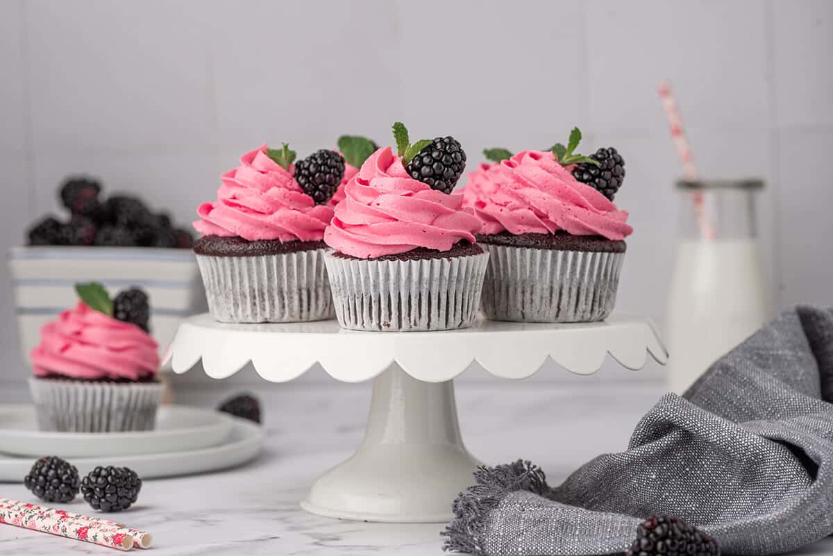 Chocolate cupcakes with blackberry buttercream frosting on a white cake stand.