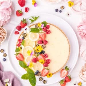 A lemon tart on a white platter topped with berries and edible flowers.
