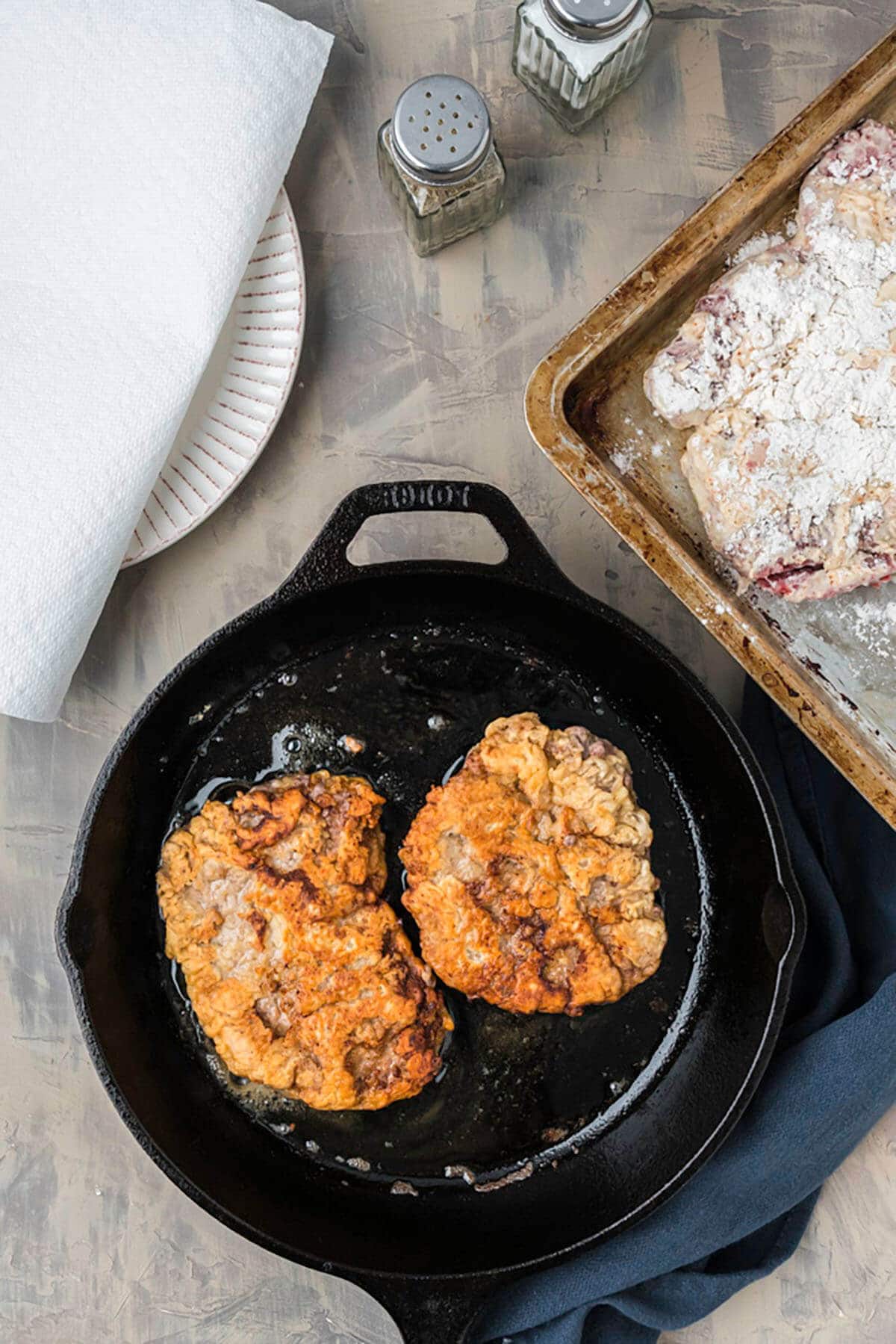 Two pieces of battered steak frying in a skillet.