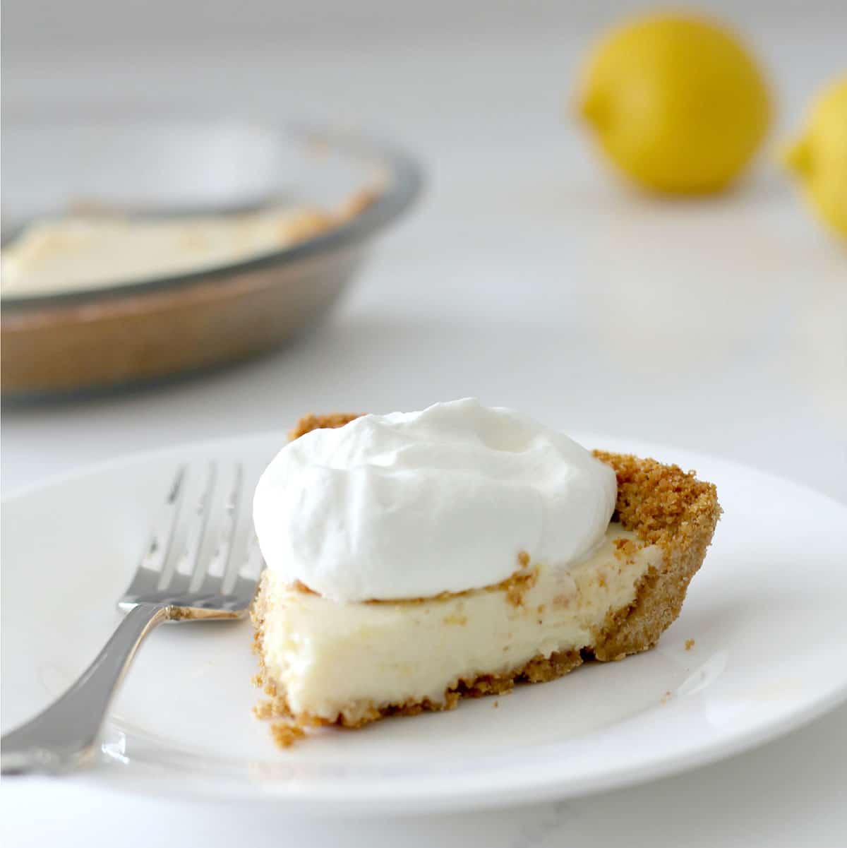 A slice of lemon icebox pie topped with whipped cream on a white plate with a fork, whole lemons, and a pie dish in the background.