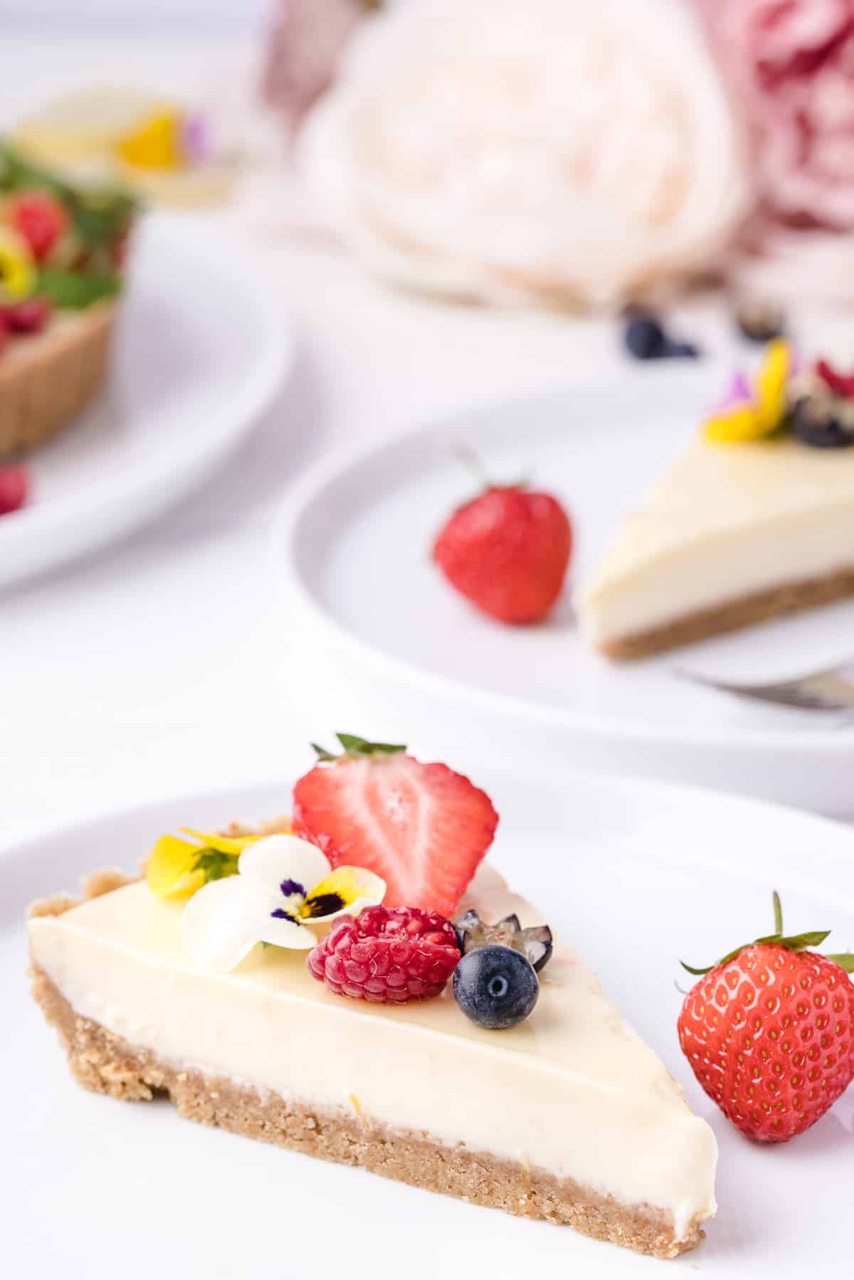 A slice of lemon icebox pie adorned with fresh berries and edible flowers, with more desserts in the background.