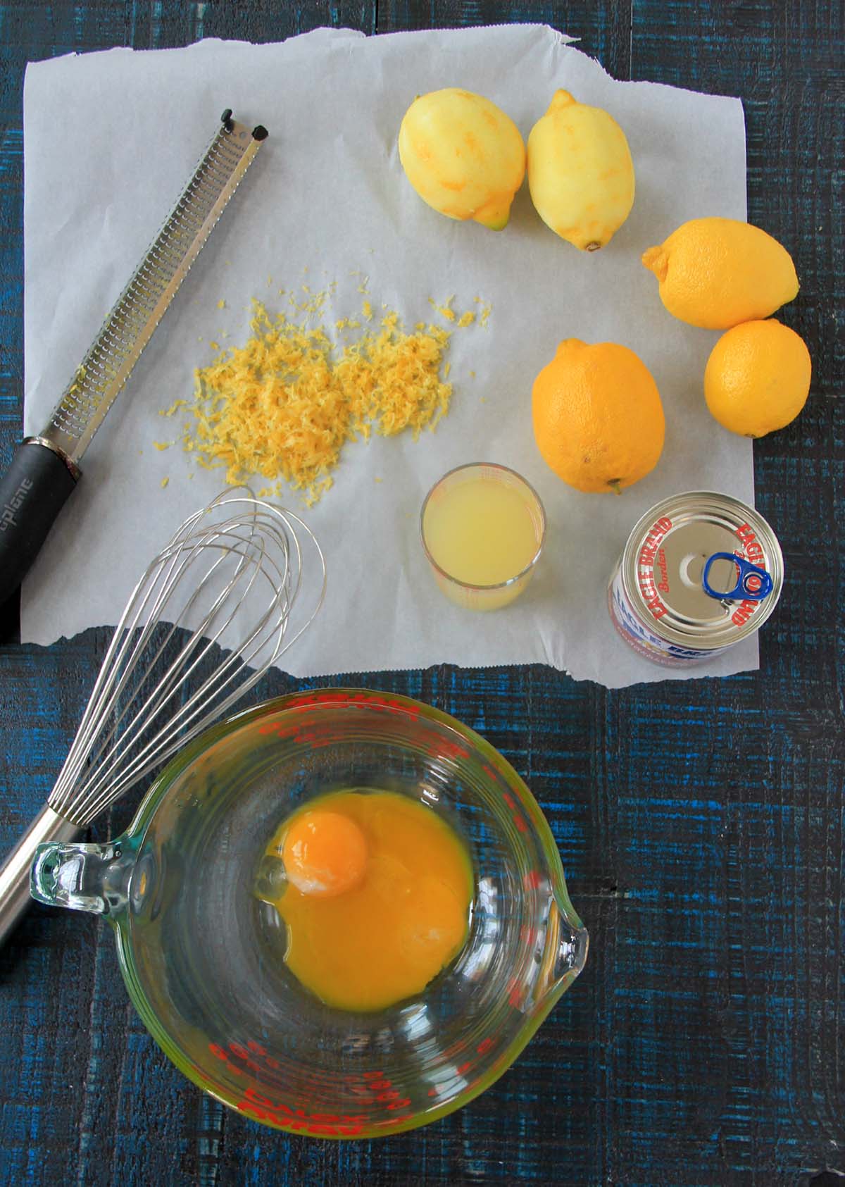 Ingredients for a lemon-based recipe arranged on a blue tablecloth, including lemons, zest, juice, an egg, and a whisk.