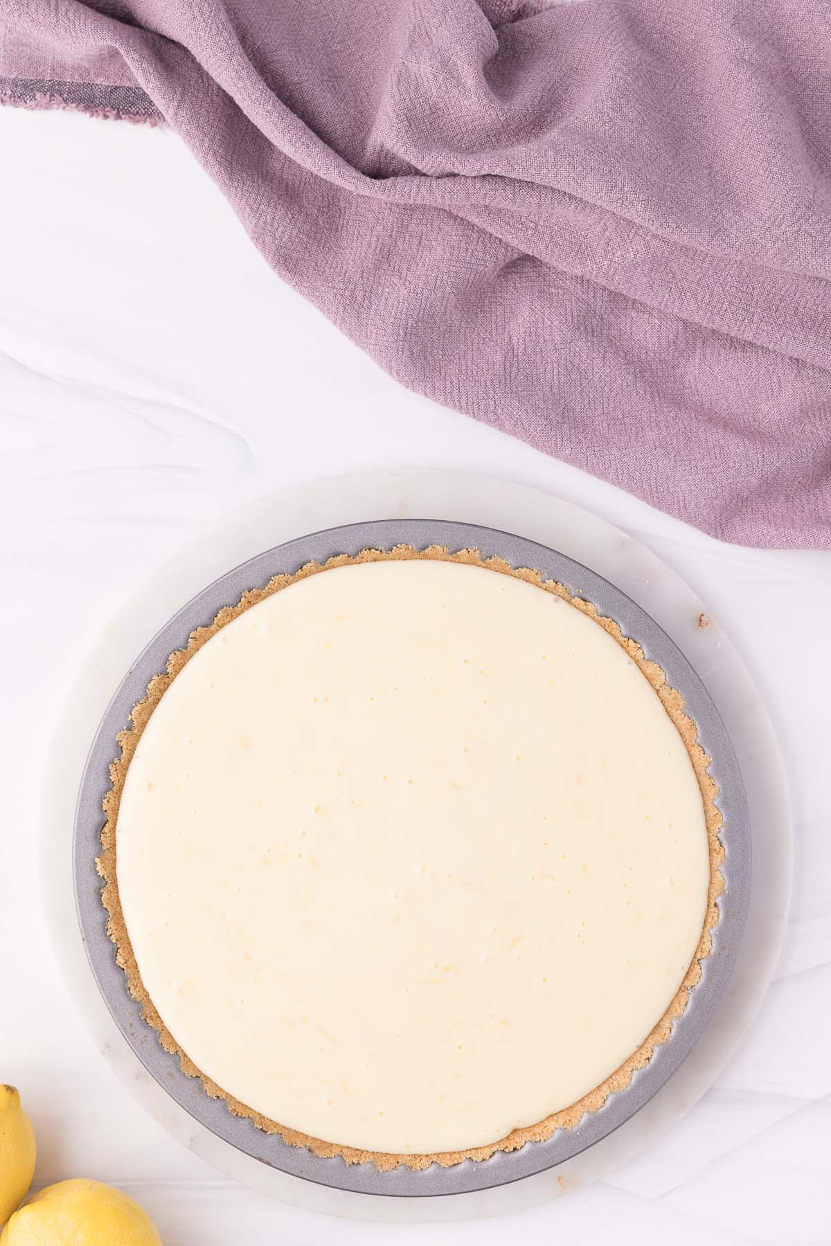 A freshly made lemon icebox pie on a gray round dish, accompanied by a purple cloth and lemons on a white surface.
