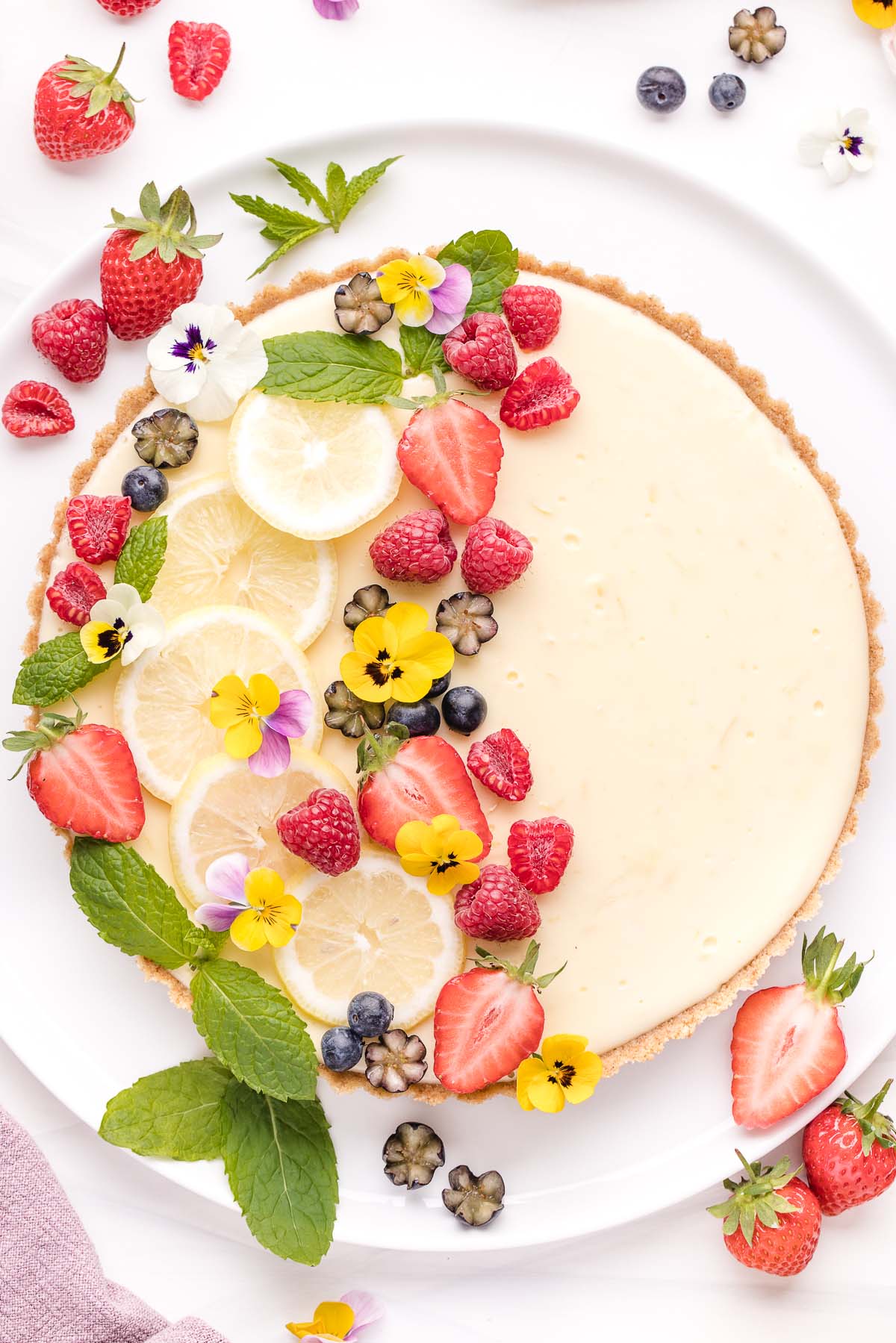 A citrus tart topped with fresh berries, lemon slices, and edible flowers on a white background.
