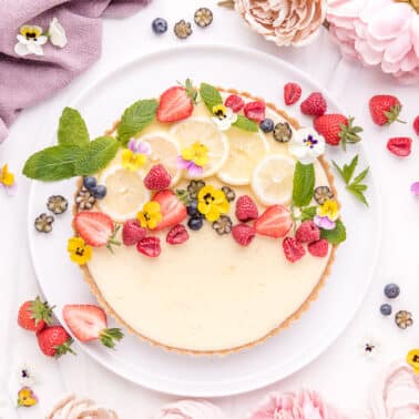 A vibrant cheesecake adorned with fresh berries, lemon slices, and edible flowers, presented on a white background with decorative elements.