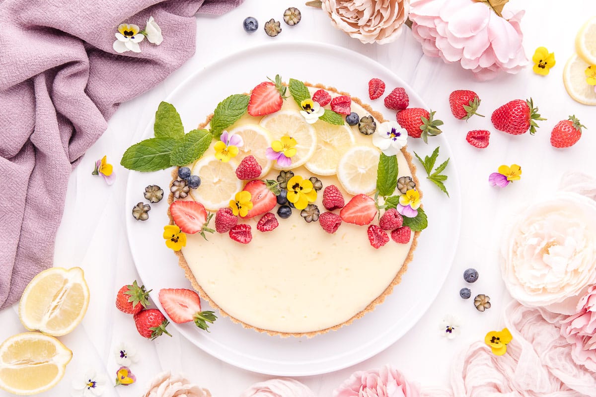 A vibrant cheesecake adorned with fresh berries, lemon slices, and edible flowers, presented on a white background with decorative elements.