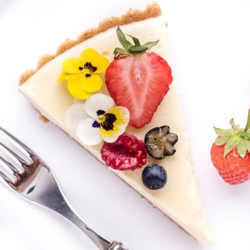 A slice of cheesecake garnished with fresh strawberries, edible flowers, and a blueberry, served with a fork on a white background.