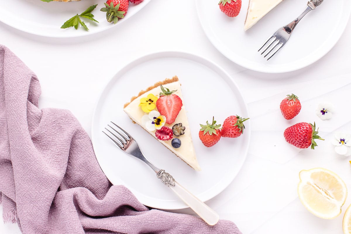 A slice of fruit tart on a white plate with strawberries and floral garnishes on a table, accompanied by a purple cloth napkin and silverware.