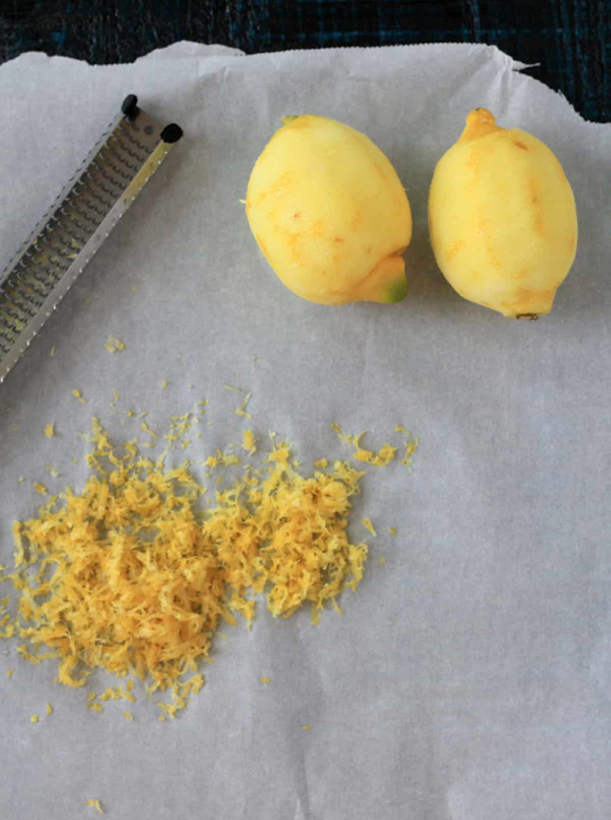 Two peeled lemons next to a pile of lemon zest and a grater on a parchment paper.