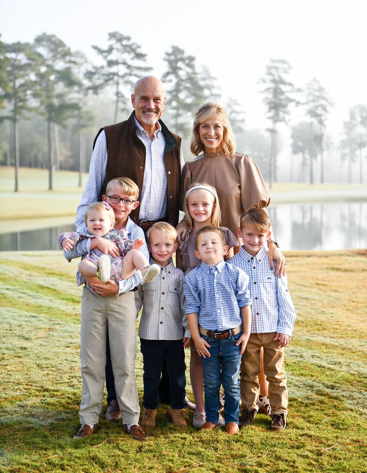 A family portrait with two adults and five young children standing outside on a grassy area with a misty lake in the background.