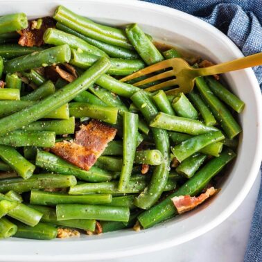 A dish of green beans seasoned with herbs and pieces of bacon, served with a fork.