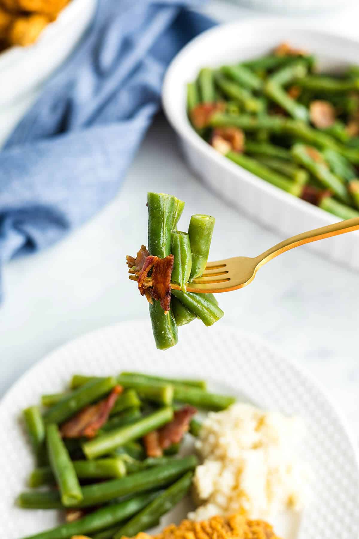 A fork holding green beans with pieces of bacon against a background of dishes with more green beans and other food.
