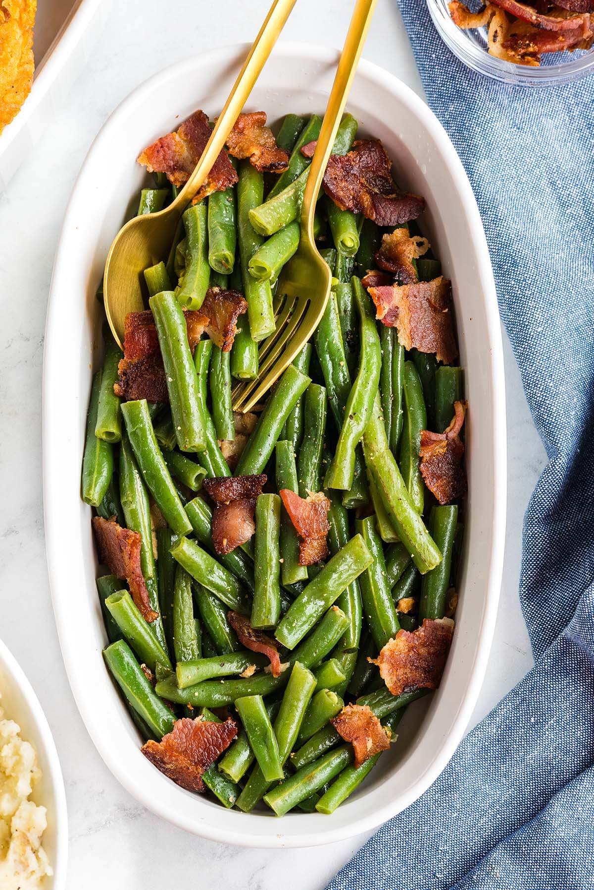A dish of cooked green beans topped with bacon pieces, served with chopsticks.