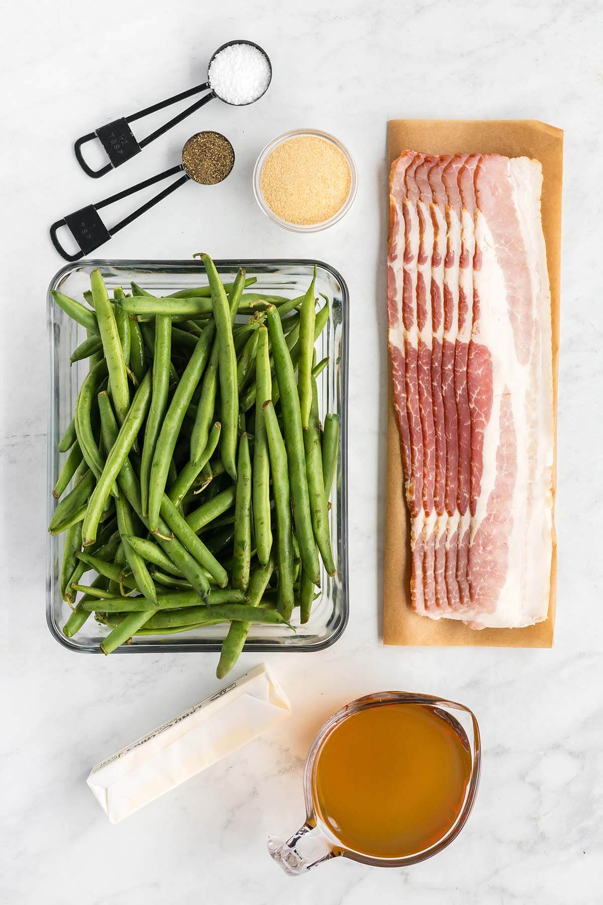 Ingredients for cooking arranged on a kitchen counter, including fresh green beans, bacon strips, seasonings, butter, and broth.