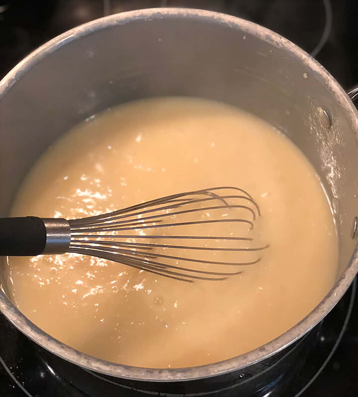 A whisk in a pot of creamy sauce cooking on a stove.