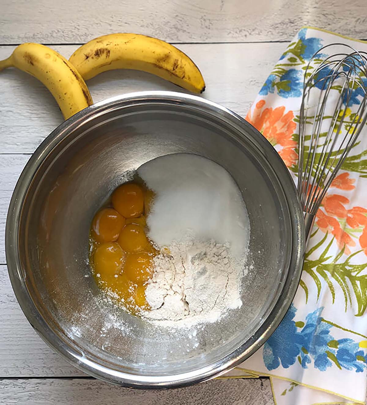A stainless steel mixing bowl containing eggs, flour, and sugar, with a whisk and two bananas beside it on a floral cloth-covered table.