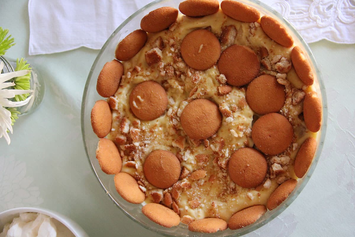 A bowl of banana pudding topped with vanilla wafers and crushed cookies, viewed from above on a light tablecloth.