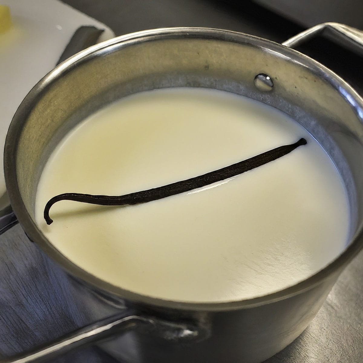 A single vanilla bean pod is steeping in a pot of milk on a stove, visible steam rising from the warm surface.