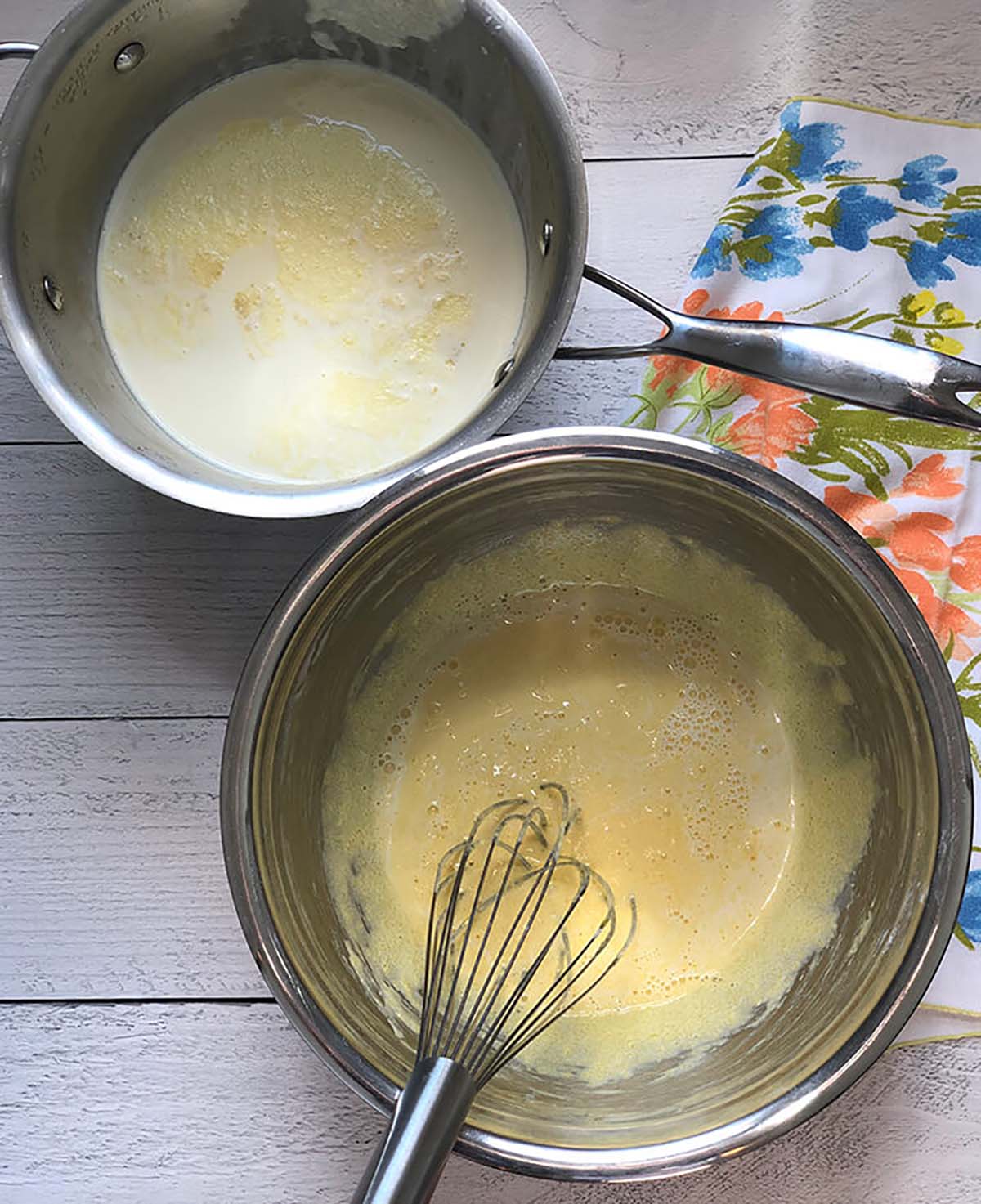 Two metal bowls on a wooden surface, one with whisked eggs and the other with milk, beside a floral cloth and a whisk.