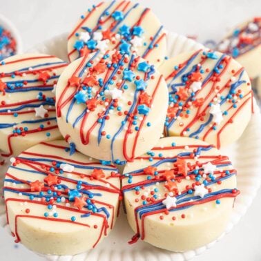 A plate of round white chocolate-covered treats decorated with red, white, and blue icing and star-shaped sprinkles, set on a white surface with a bowl of matching sprinkles nearby.