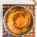 Peach barbecue sauce simmering in a circular metal pan, placed on a wooden trivet, with a blue cloth to the side and a wooden spoon in the top right corner.