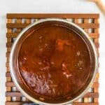 A pot of thick peach bourbon barbecue sauce with a wooden spoon placed next to it on a wooden trivet.
