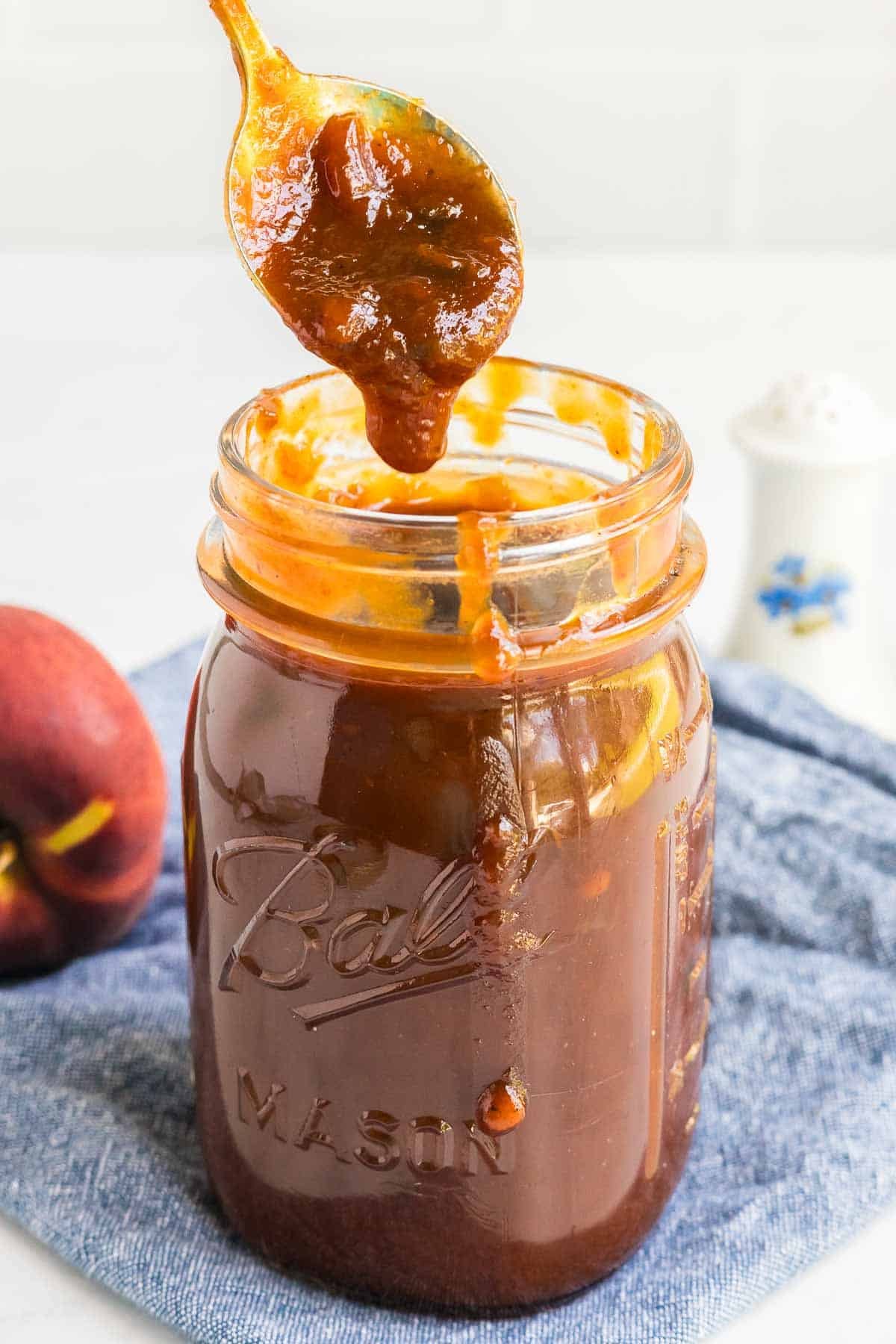 A spoon dipped in dark peach BBQ sauce hovers over a mason jar filled with the same sauce. A fresh peach is partially visible in the background.