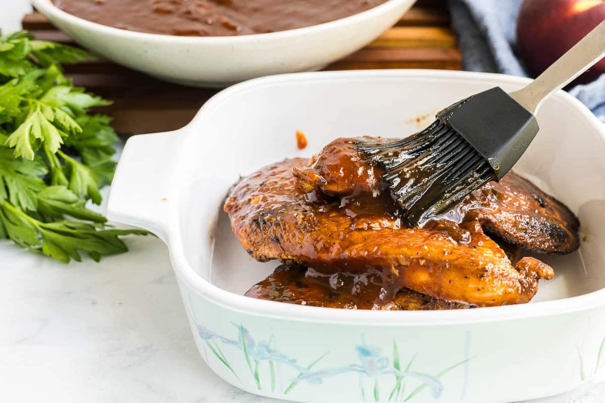A brush applies barbecue sauce to cooked chicken breasts in a white ceramic dish. Fresh parsley and a bowl of more sauce are in the background.