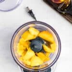 A food processor with chopped peaches inside sits on a countertop. A partially peeled peach and a peeler are on a cutting board beside it.