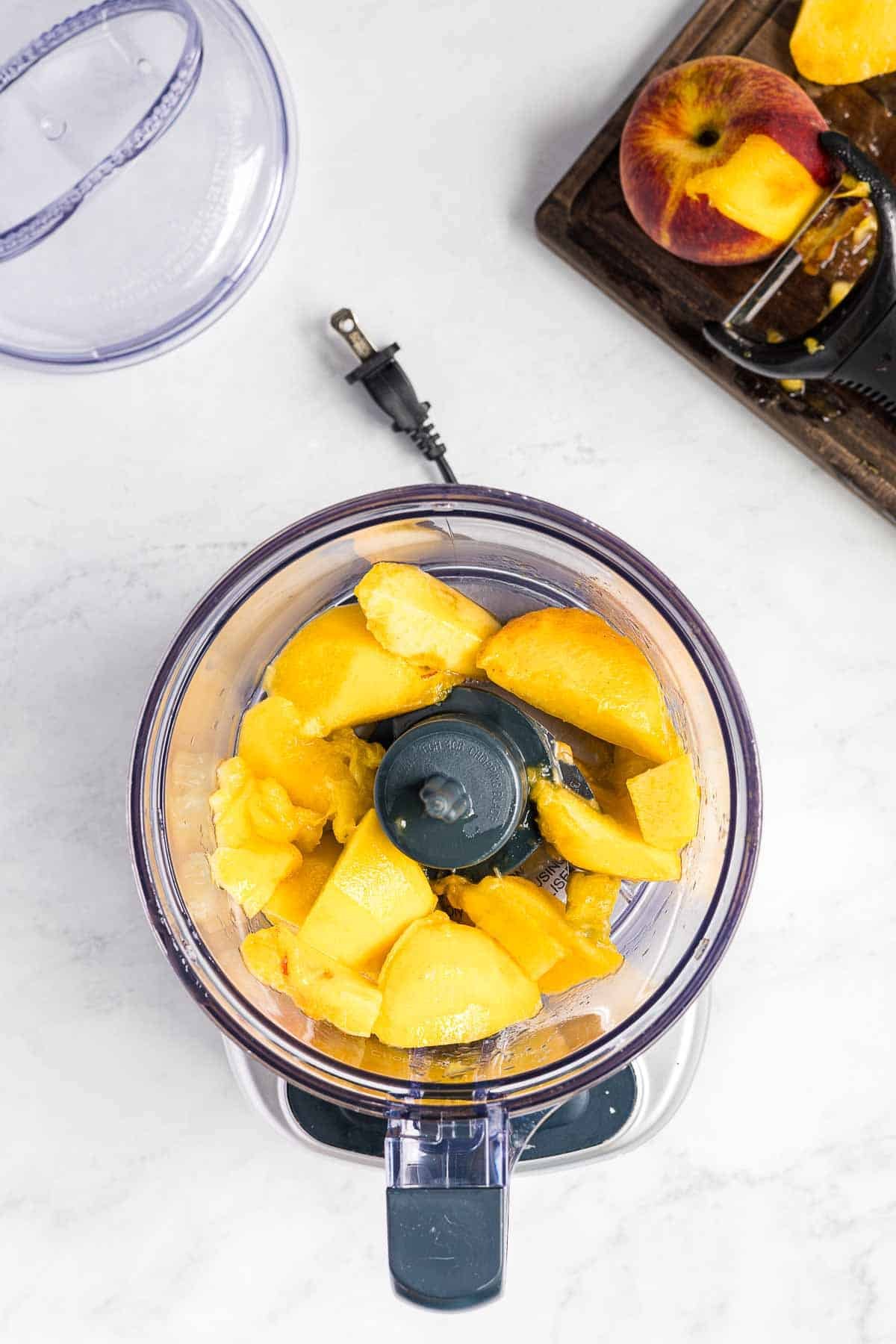 A food processor with chopped peaches inside sits on a countertop. A partially peeled peach and a peeler are on a cutting board beside it.