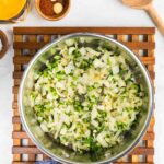 A stainless steel pot filled with diced onions and green peppers rests on a wooden trivet. Various ingredients and a wooden spoon are placed nearby.