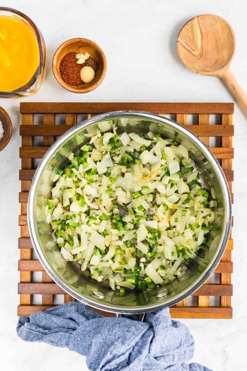 A stainless steel pot filled with diced onions and green peppers rests on a wooden trivet. Various ingredients and a wooden spoon are placed nearby.