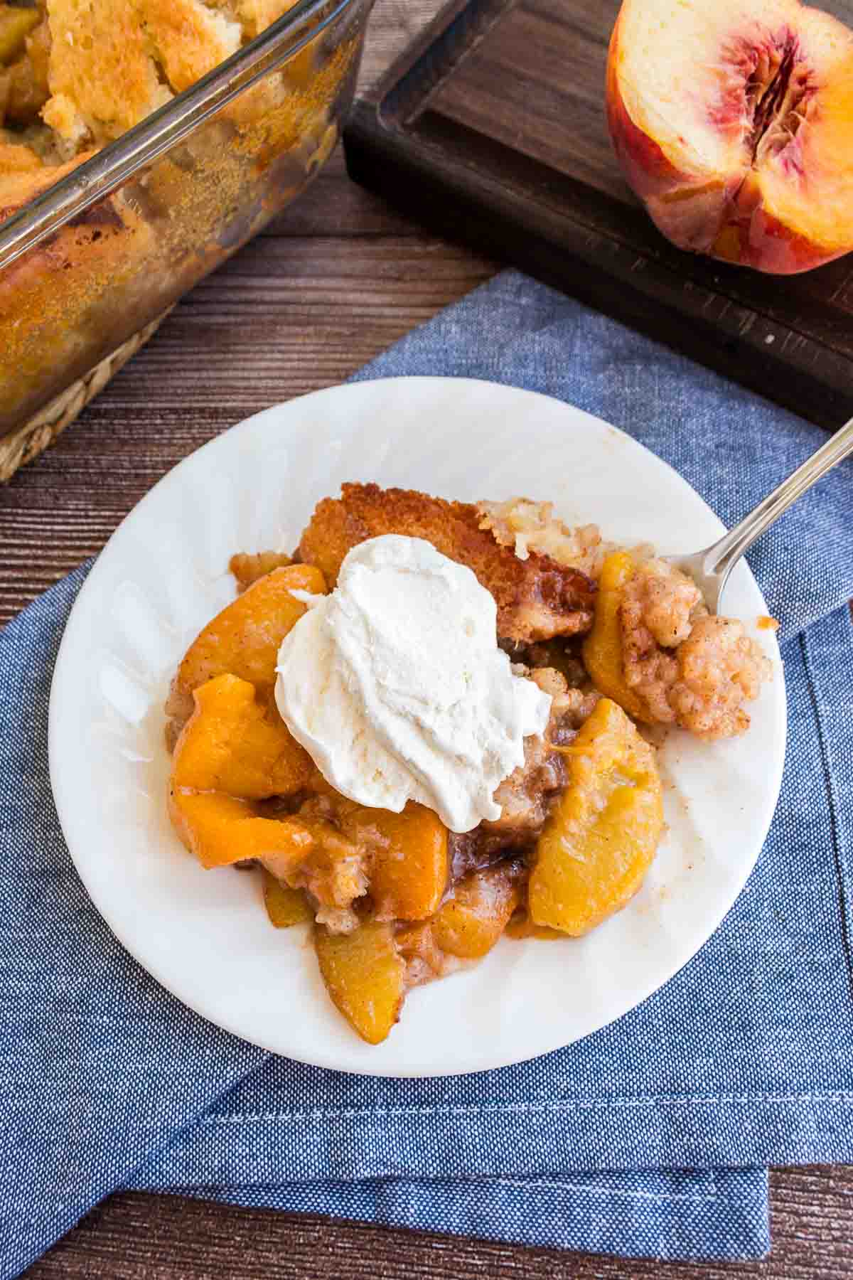 A plate of peach cobbler topped with a dollop of whipped cream sits on a blue napkin, with a partially sliced peach and a baking dish of cobbler in the background.