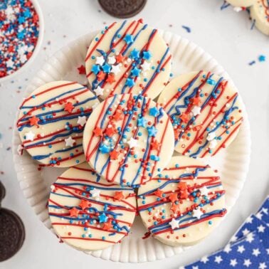 A plate of round white chocolate-covered Oreos decorated with red, white, and blue sprinkles and drizzle, arranged on a white dish with additional sprinkles and Oreos in the background.