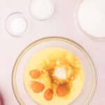 A baking preparation setup with raw eggs, flour, sugar, and salt in separate bowls on a pink surface.