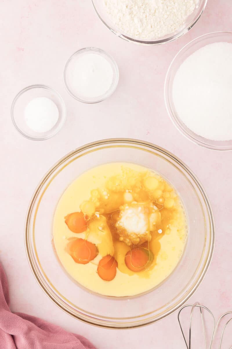 A baking preparation setup with raw eggs, flour, sugar, and salt in separate bowls on a pink surface.