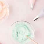 Overhead view of three bowls of pastel-colored frosting with piping bags on a pink surface.