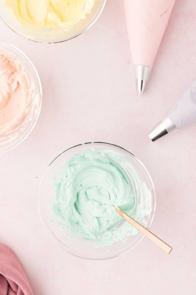 Overhead view of three bowls of pastel-colored frosting with piping bags on a pink surface.