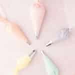 Five piping bags with different colored icings (pink, peach, lavender, yellow, and mint) positioned on a light pink background.