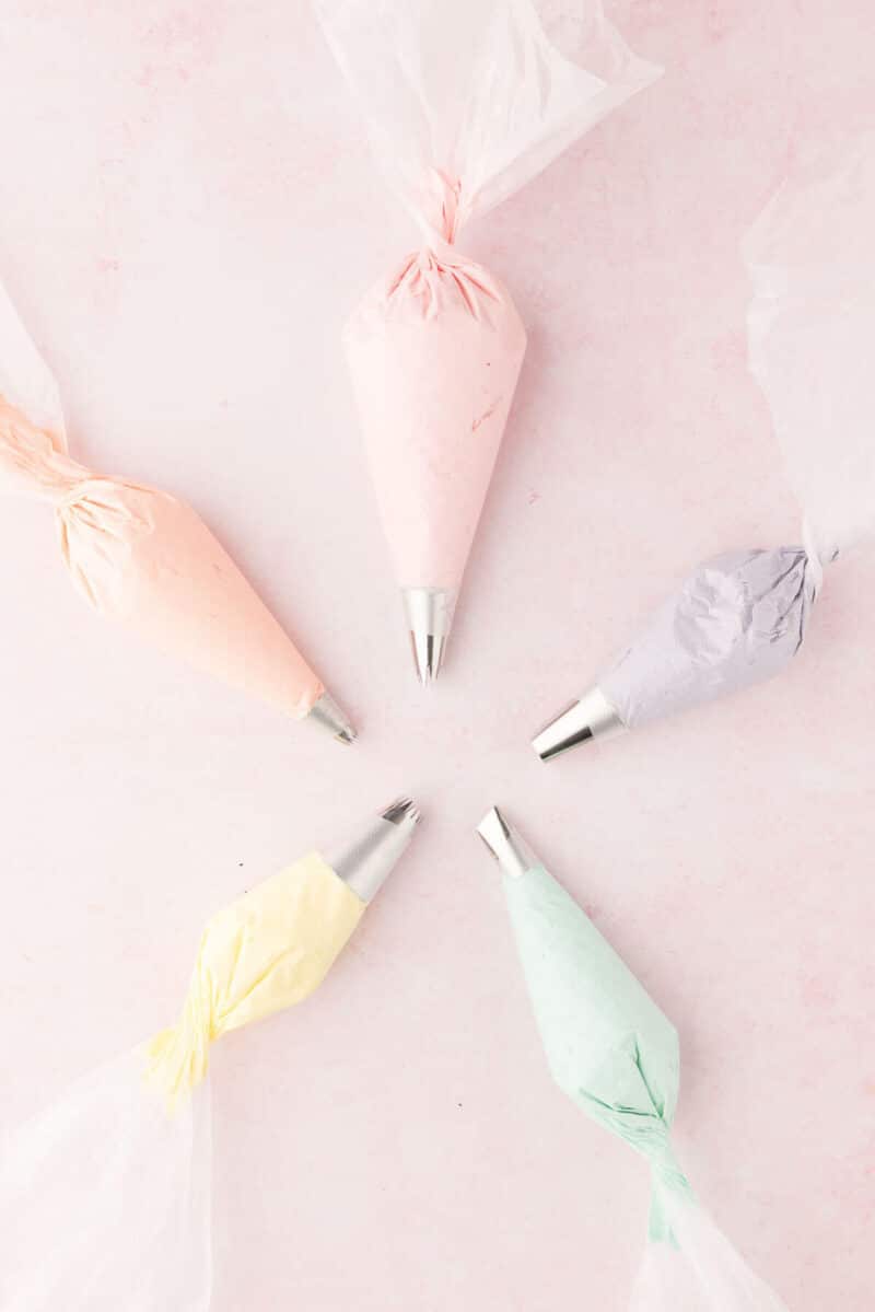 Five piping bags with different colored icings (pink, peach, lavender, yellow, and mint) positioned on a light pink background.