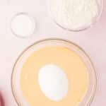 Glass bowl with beaten eggs and sugar on a pink surface, next to bowls of flour and salt, ingredients prepared for baking.