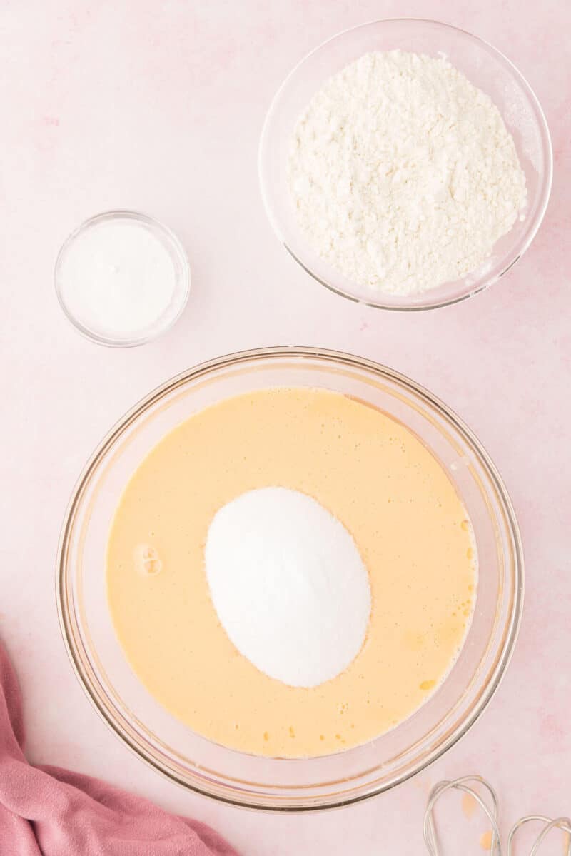 Glass bowl with beaten eggs and sugar on a pink surface, next to bowls of flour and salt, ingredients prepared for baking.