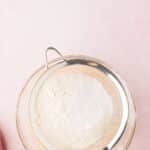 Flour being sifted through a metal sieve into a pink bowl on a pale pink surface, with a person holding the sieve.
