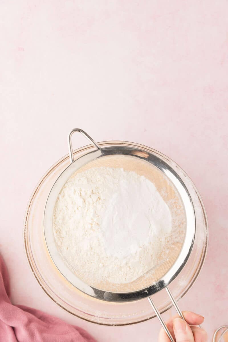 Flour being sifted through a metal sieve into a pink bowl on a pale pink surface, with a person holding the sieve.