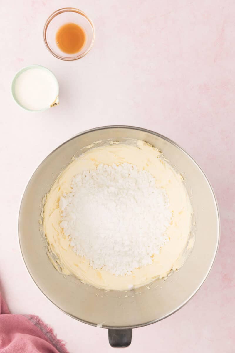 A mixing bowl with butter and sugar creamed together, with a separate glass of milk and a shot of vanilla extract on a pink surface.