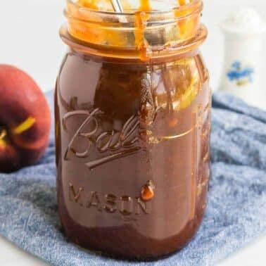 A glass Mason jar filled with sauce, placed on a blue cloth with a peach partially visible in the background.