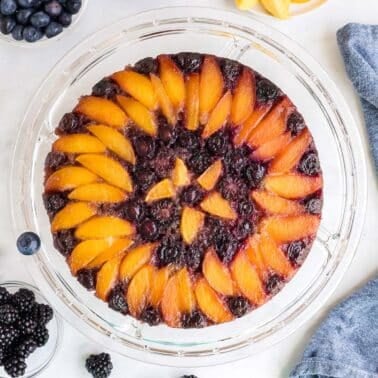 A round peach upside down cake on a glass plate with peach slices and blueberries arranged in concentric circles. Several small bowls of blackberries and blueberries are around the cake.
