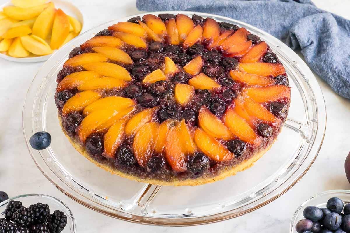 A peach blueberry cake on a glass plate, topped with sliced peaches and blueberries arranged in a circular pattern. Bowls with additional fruit are visible in the background.