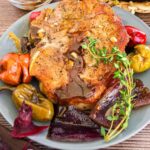 Pork chop on a plate with assorted vegetables and a drizzle of sauce, garnished with fresh herbs.