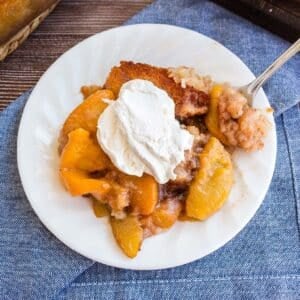 A white plate holds a serving of peach cobbler topped with a dollop of whipped cream. A silver fork is placed on the plate, resting on top of a blue cloth napkin.